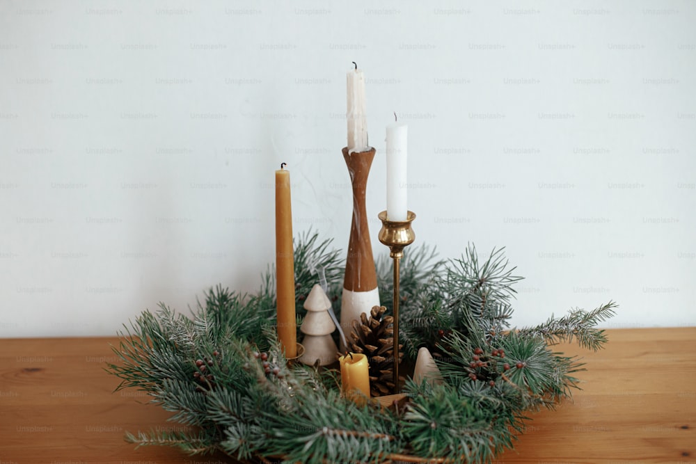 Stylish candles in christmas wreath  with pine cones and tree decor on wooden table on background of white wall in festive room. Christmas advent. Atmospheric winter holidays time