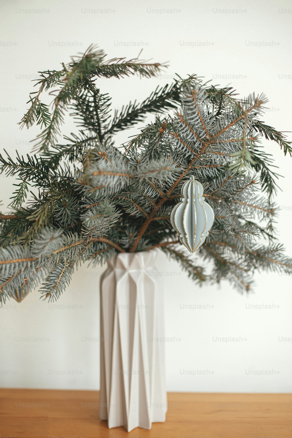 Merry Christmas! Stylish christmas ornament on fir branch in modern vase close up. Modern wooden bauble on branch on white background. Festive simple decor in scandinavian room.