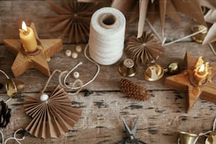 素朴な木製の背景にスタイリッシュなクリスマスの鐘、紙の天使と星、松ぼっくり、ろうそく、糸、はさみ。お祝いのスカンジナビアの花輪、不機嫌なイメージを作ります。手作りの休日の装飾