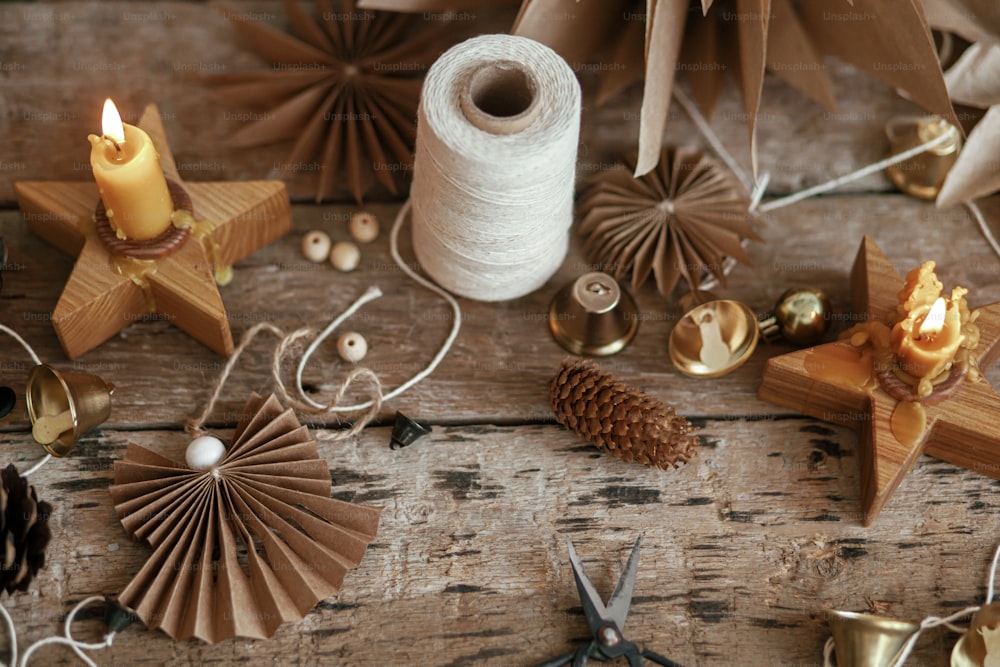 Stylish Christmas bells, paper angel and stars, pine cones, candle, thread and scissors on rustic wooden background. Making festive scandinavian garland, moody image. Handmade holiday decor