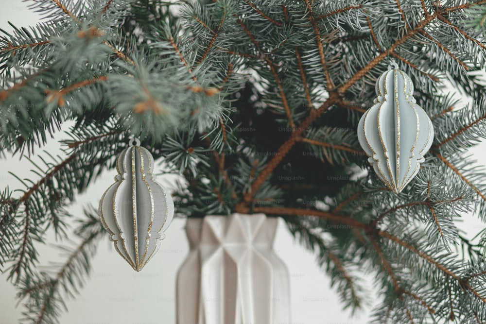 Stylish christmas ornament on fir branch close up. Modern wooden  bauble on branch in modern grey vase on white background. Festive decor in modern scandinavian room. Merry Christmas!