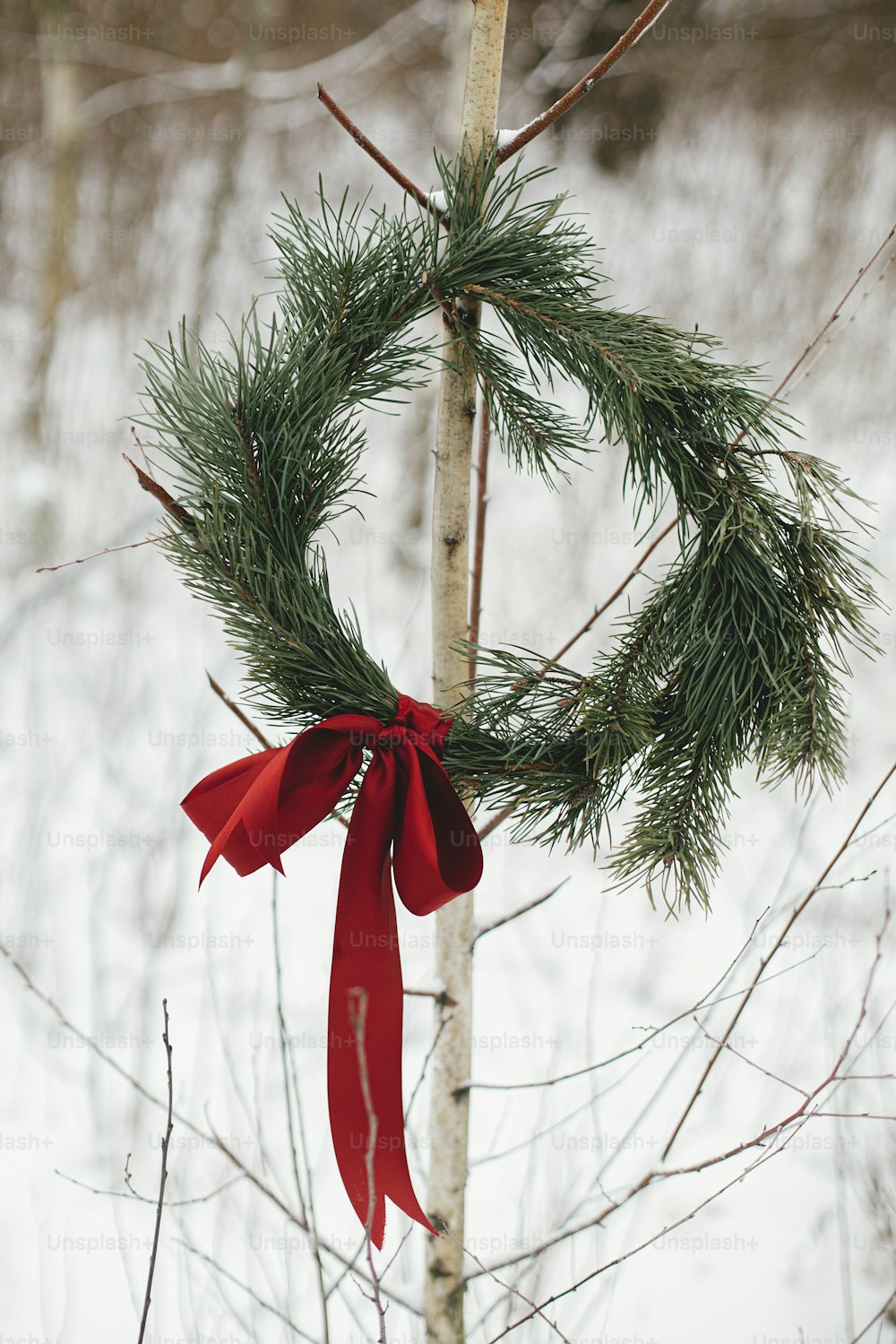 Stilvoller Weihnachtskranz am Baum im verschneiten Winterpark. Einfacher Weihnachtskranz mit Tannenzweigen und roter Schleife im Freien. Rustikale Öko-Dekoration. Urlaub auf dem Land. Frohe Weihnachten!