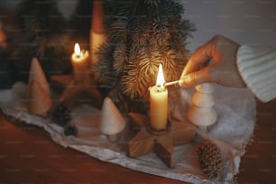 Holiday advent. Hand in cozy sweater lighting up christmas candle on background of pine trees decorations, cones, rustic cloth on wooden table in evening scandinavian festive room.  Atmospheric