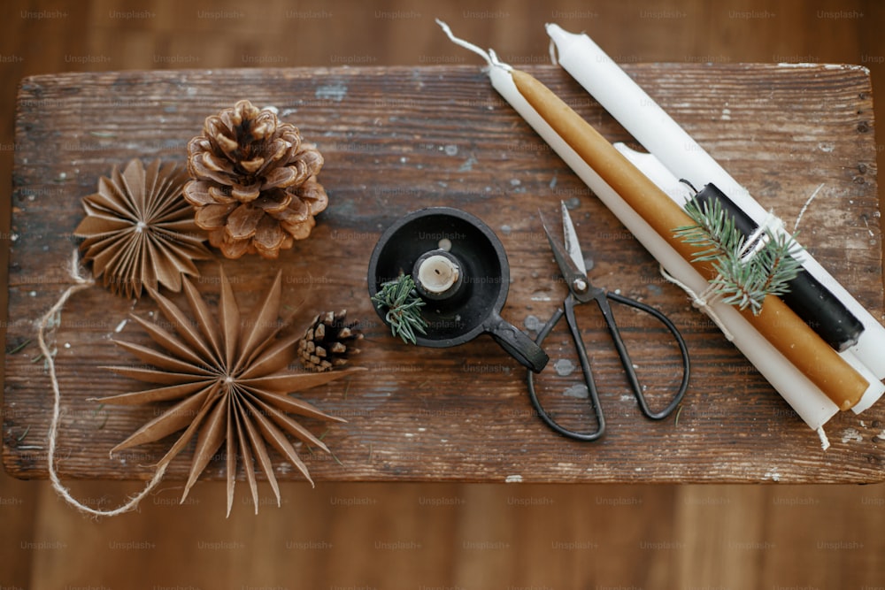 Stylish Christmas candles, paper stars, pine cones and fir branch, scissors on rustic wooden background. Top view. Festive holiday decor. Atmospheric Holiday advent. Scandinavian hygge