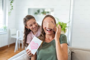 Happy little girl congratulating smiling mother and giving card with red heart during holiday celebration at home, covering her eyes and surprising her