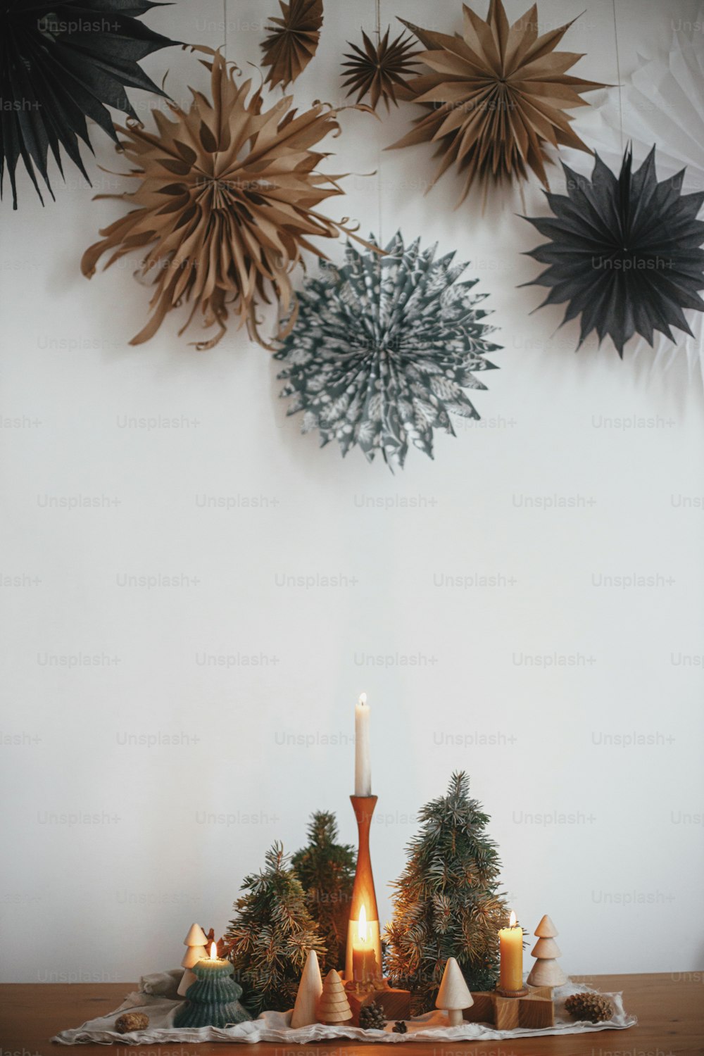Stylish christmas candles and trees decorations on wooden table on background of white wall with big paper stars. Handmade holiday decor. Atmospheric winter time. Merry Christmas!