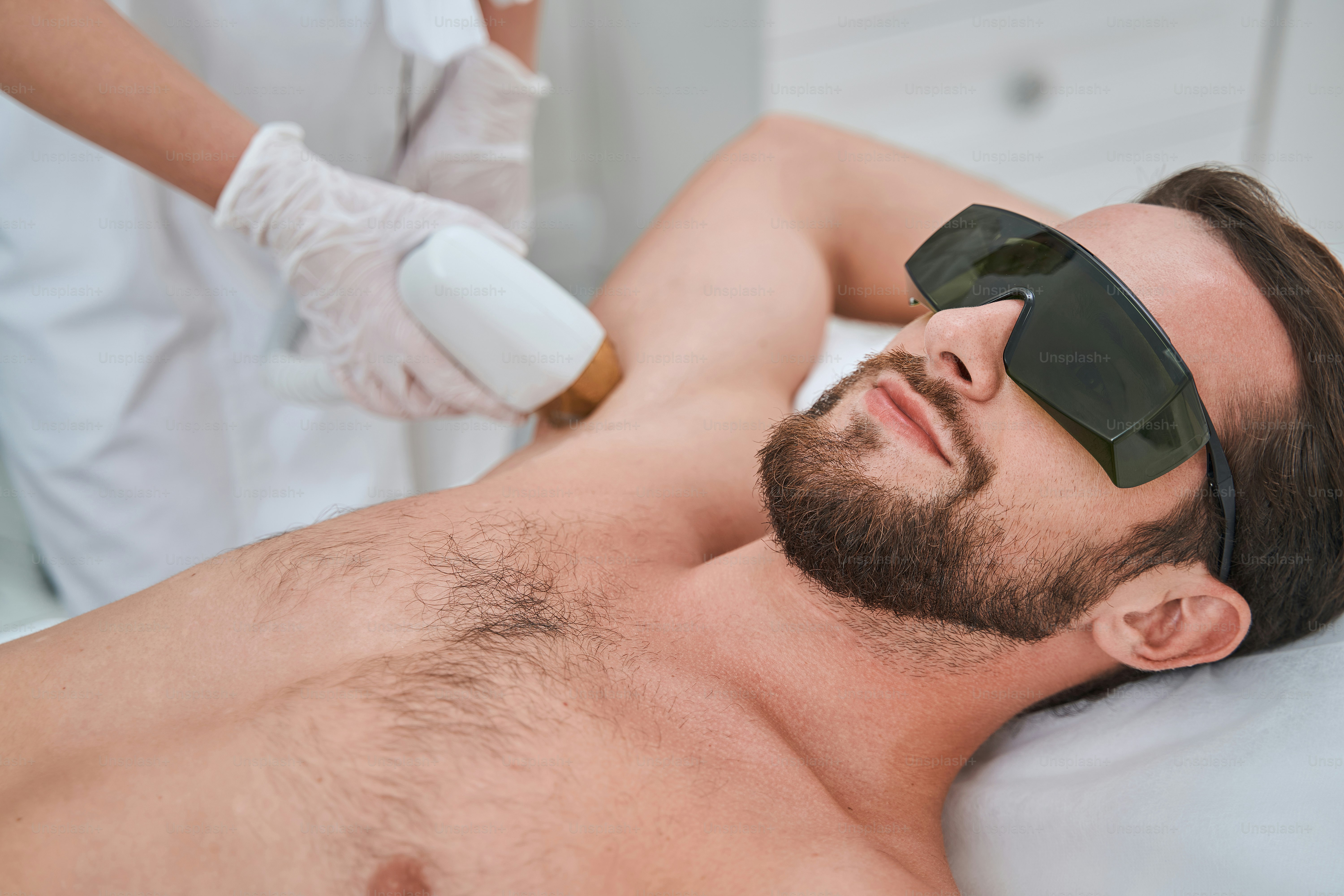 Homme barbu aux cheveux noirs portant des lunettes de protection subissant une épilation au laser des aisselles dans un salon de beauté