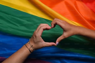Hand of LGBT women holding together and forming heart shape over rainbow flag. Concept of LGBT pride.