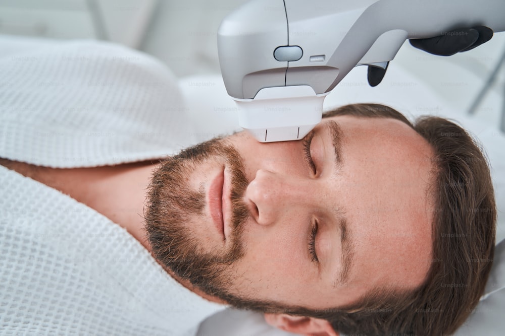 Handsome male patient lying with his eyes shut during the ultrasound therapy performed by a cosmetologist