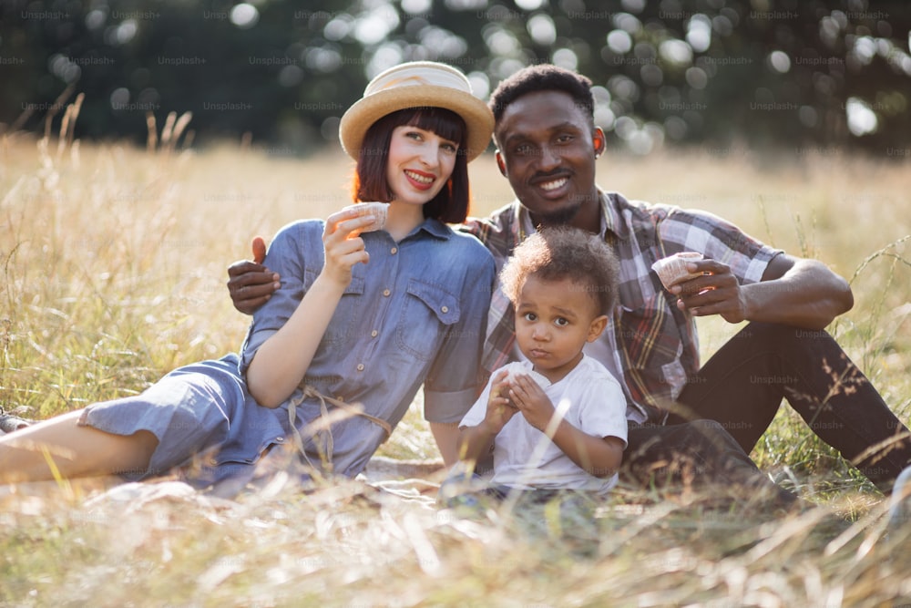 Portrait of beautiful mixed race parents sitting with little son on grass and eating tasty cupcakes. Happy family of three enjoying summer picnic on fresh air.