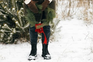 Stylish woman with christmas wreath in winter park. Young female in cozy mittens and coat holding xmas wreath with red bow in snowy winter field, cropped view. Merry Christmas!