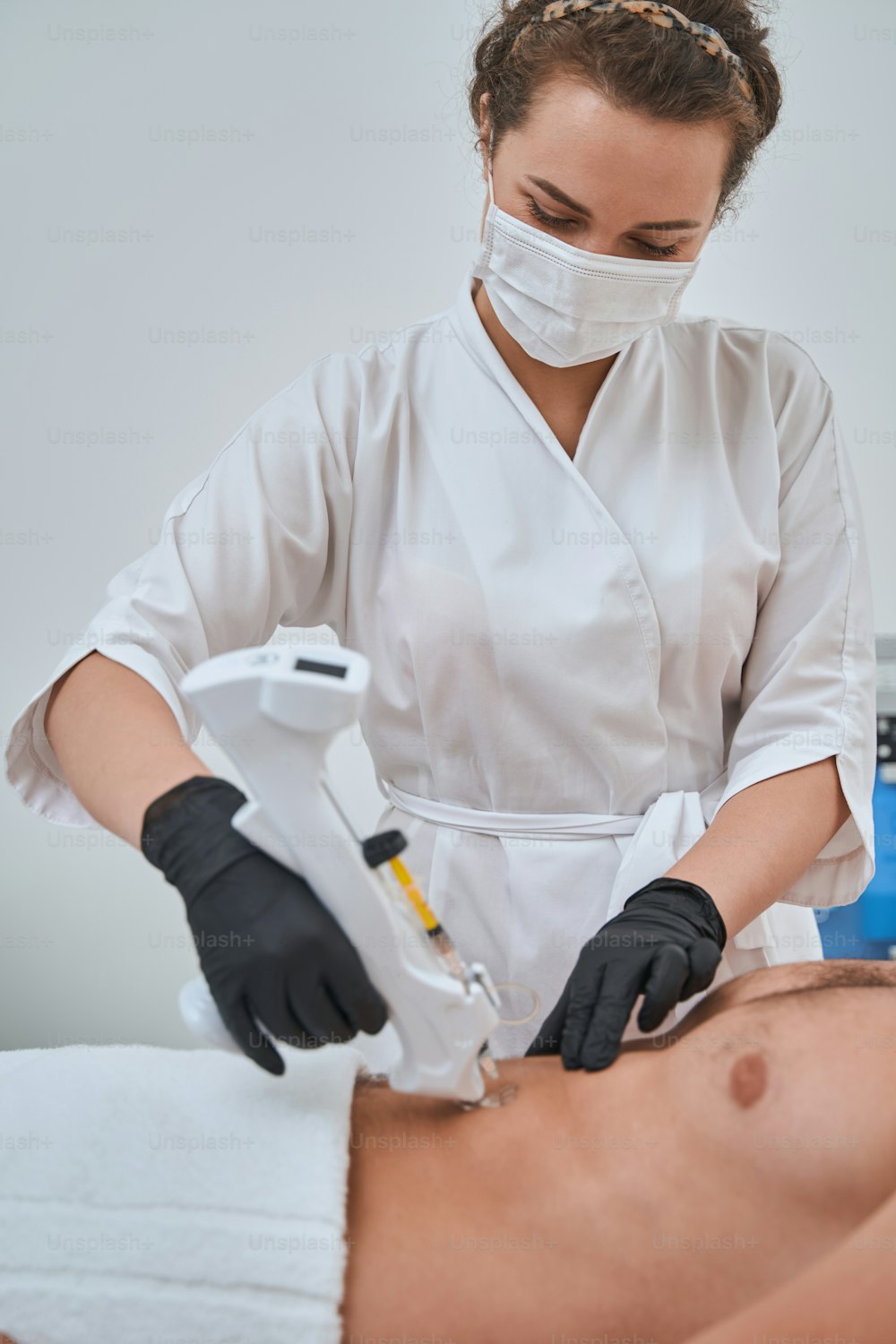 Focused dermatologist in a face mask and nitrile gloves injecting a male patient into the abdomen with an injection device
