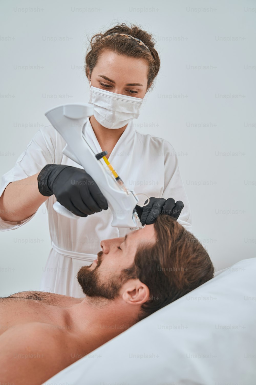 Focused female dermatologist in nitrile gloves injecting a dermal filler into a male patient forehead using a mesogun