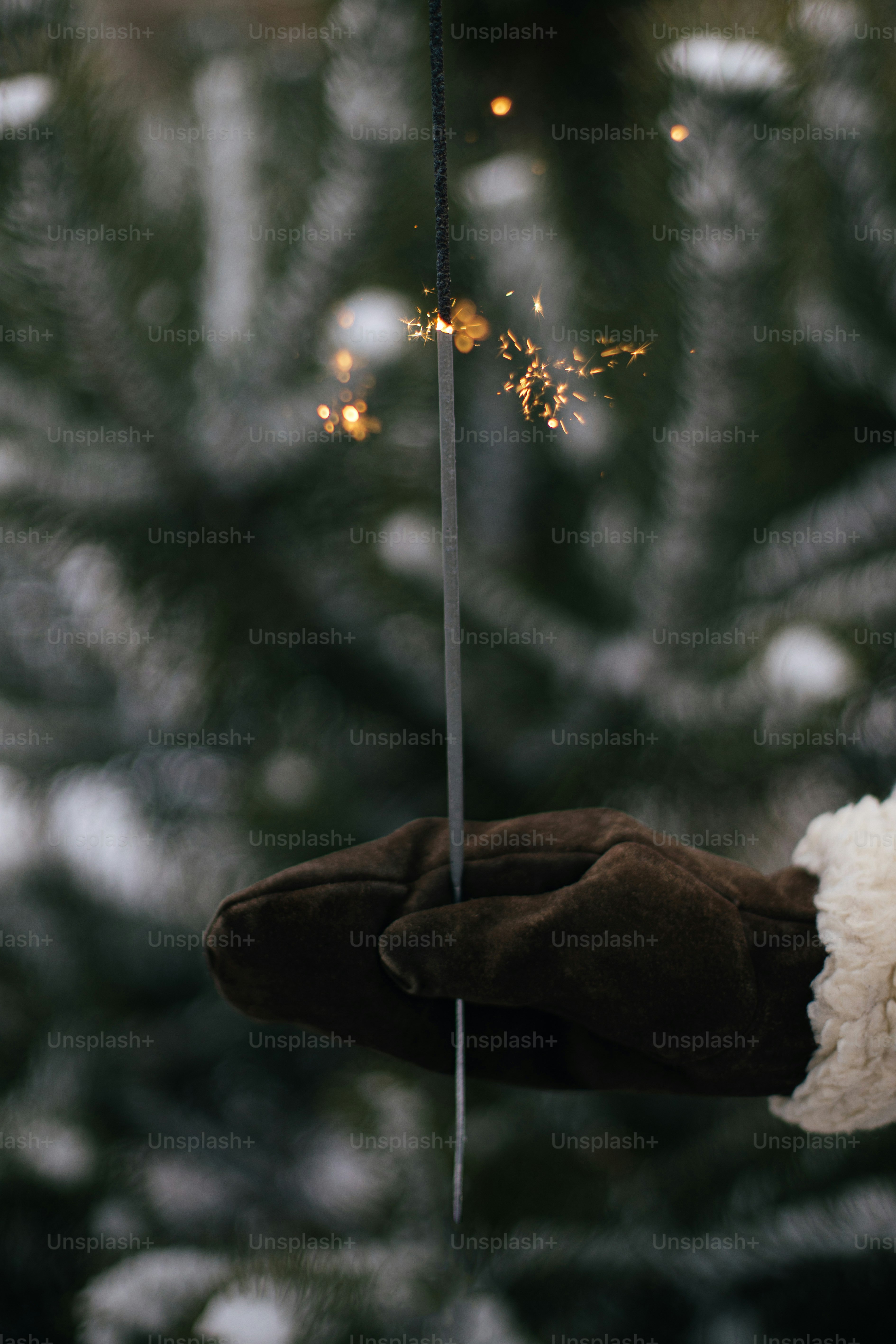 Happy New Year!  Woman hand in stylish mitten with glowing firework on background of pine tree branches in snow in evening. Hand holding burning sparkler. Atmospheric magic moment. Happy Holidays!