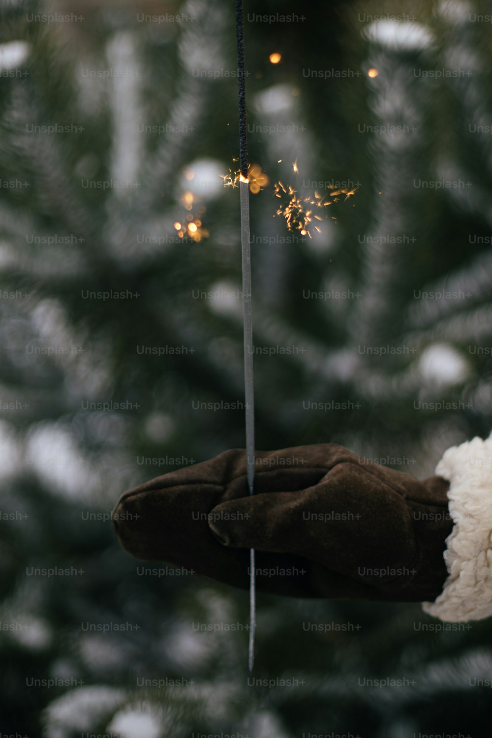 Happy New Year!  Woman hand in stylish mitten with glowing firework on background of pine tree branches in snow in evening. Hand holding burning sparkler. Atmospheric magic moment. Happy Holidays!