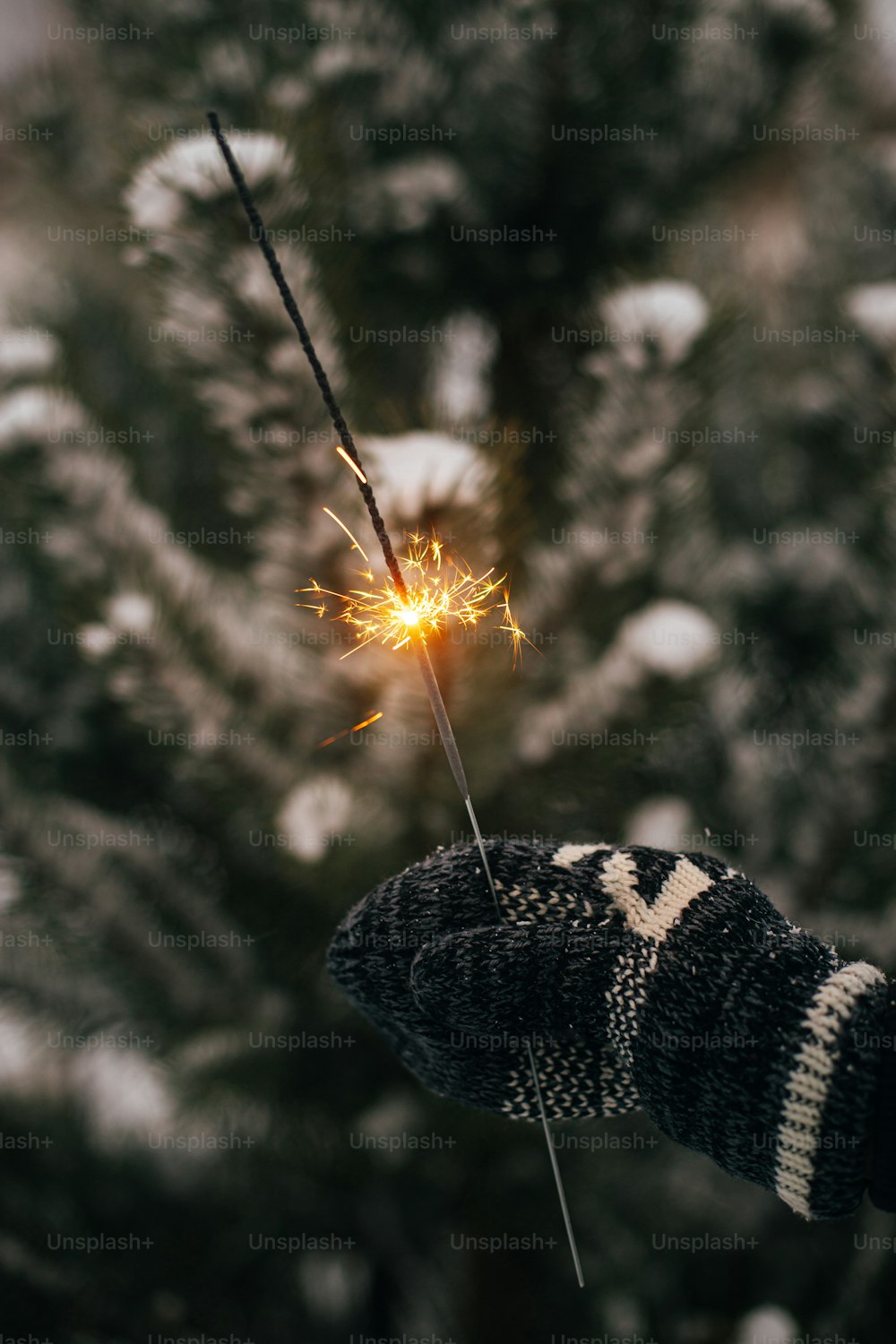 Happy New Year!  Woman hand in cozy mitten with burning firework on background of pine tree branches in snow in evening. Hand holding glowing sparkler. Atmospheric magic moment
