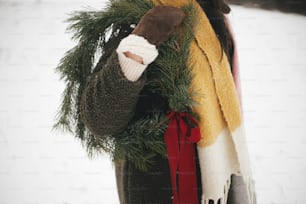 Stilvolle schöne Frau mit Weihnachtskranz im verschneiten Winterfeld. Junge Frau in kuscheligen Fäustlingen und Mantel mit Weihnachtskranz mit roter Schleife. Urlaub auf dem Land. Magische Winterzeit, beschnittene Ansicht
