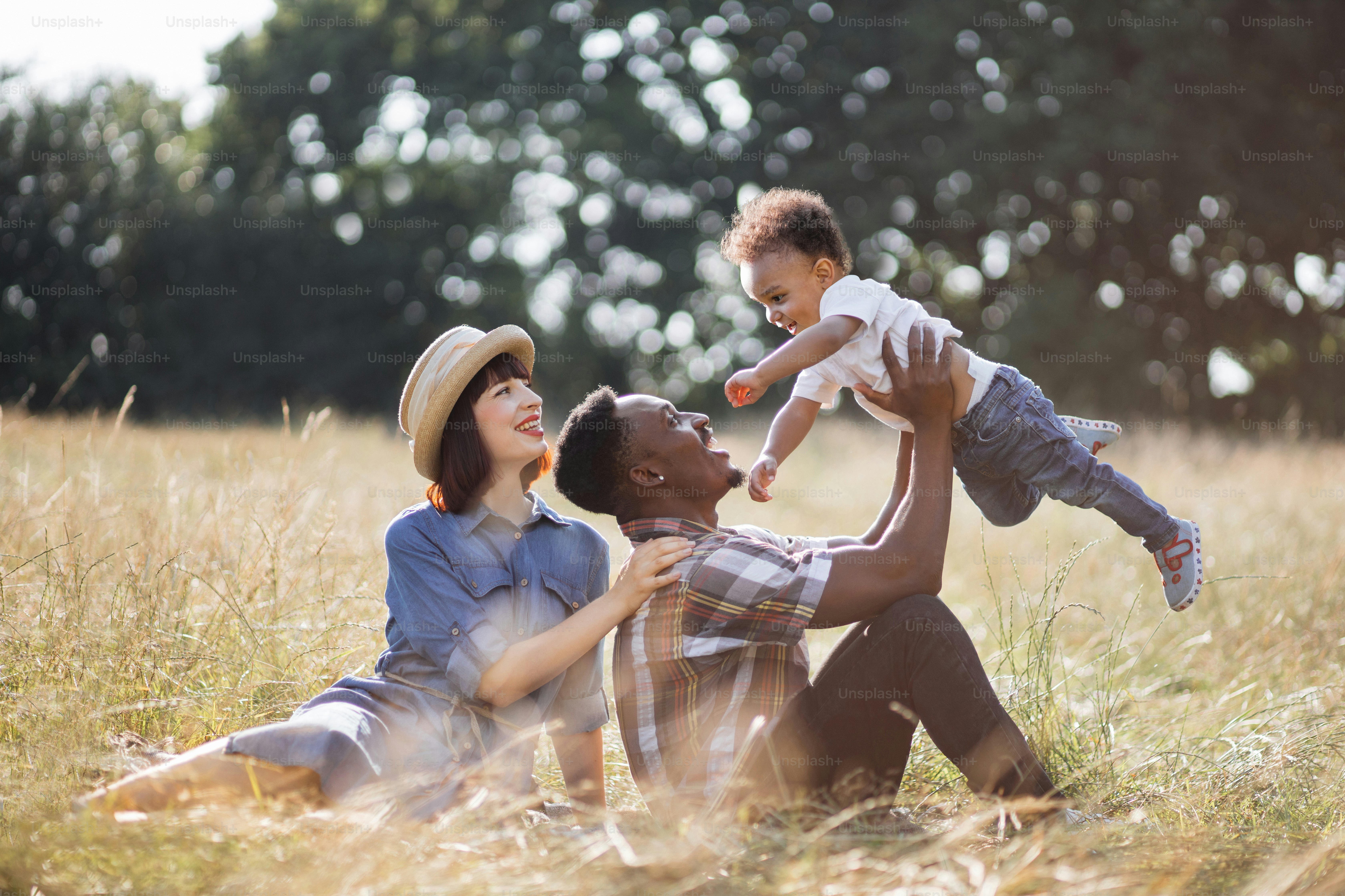 Multirassische dreiköpfige Familie, die den Sommer zum Picknick an der frischen Luft verbringt. Afrikanischer Vater spielt mit kleinem Sohn, während kaukasische Mutter in der Nähe sitzt und aufrichtig lächelt.