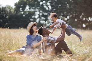 Multiracial family of three spending summer time for picnic on fresh air. African father playing with little son while caucasian mother sitting near and smiling sincerely.