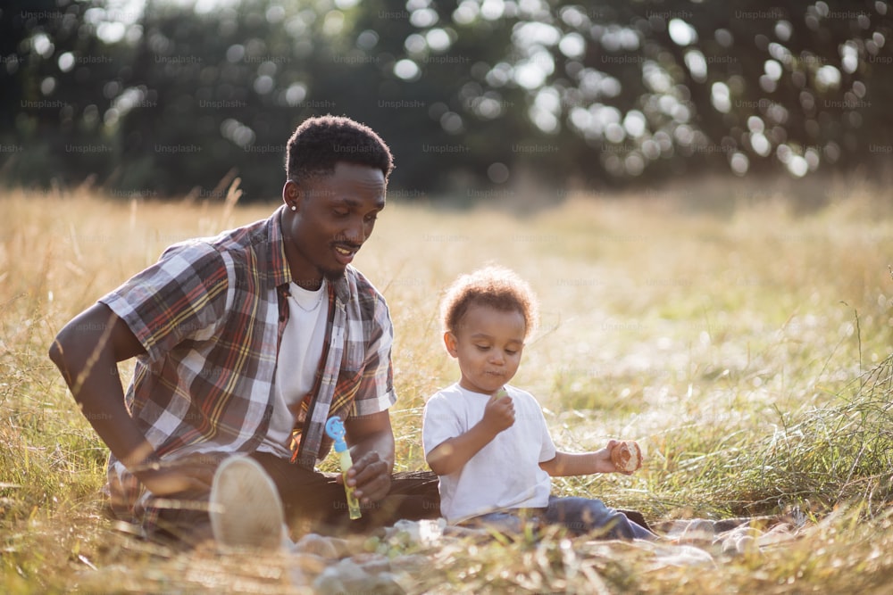 Fürsorglicher afrikanischer Vater, der mit seinem kleinen Sohn im hohen Gras sitzt und mit Seifenblasen spielt. Sommerpicknick der glücklichen Familie. Spielerische Stimmung und Entspannung.