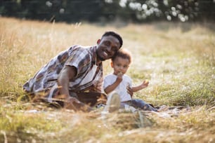 Pai afro-americano abraçando o filho pequeno e sorrindo na câmera enquanto sentados juntos no campo de verão. Menino bonito comendo uva doce durante o piquenique com o pai carinhoso.