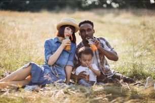 Portrait of happy multi ethnic parents embracing little cute son while sitting on wild field and drinking fresh juice. Family on picnic. Summer time concept.