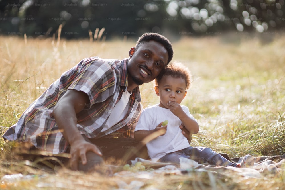 Cute little african boy eating fresh grape during summer picnic with his father. African american man gently hugging his son and smiling on camera while sitting on grass.