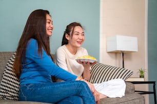 Amigas asiáticas milenarias sentadas en el sofá viendo películas en la televisión con comer papas fritas juntas en la sala de estar. Amistad femenina disfrutar y divertirse Fin de semana actividad estilo de vida con entretenimiento en el hogar