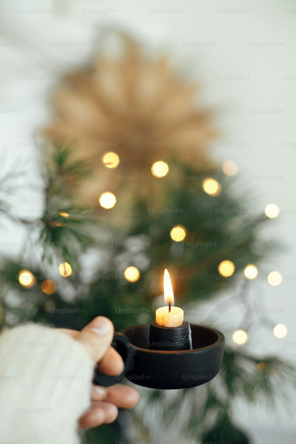 Hand in cozy sweater holding vintage candlestick with burning candle on background of warm lights, fir branches, sweden star in festive scandinavian room. Atmospheric hygge home. Winter time