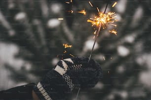 ¡Feliz Año Nuevo! Mano en guante acogedor sosteniendo bengala encendida en el fondo de ramas de pino en la nieve. Momento mágico atmosférico. Mano de mujer con fuegos artificiales brillantes en la noche. ¡Felices Fiestas!