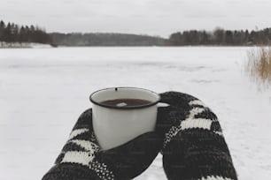 Hands in cozy gloves holding warm cup of tea on background of  snow lake in winter. Hiking and traveling in cold winter season. Warm drink. Space for text. Wanderlust