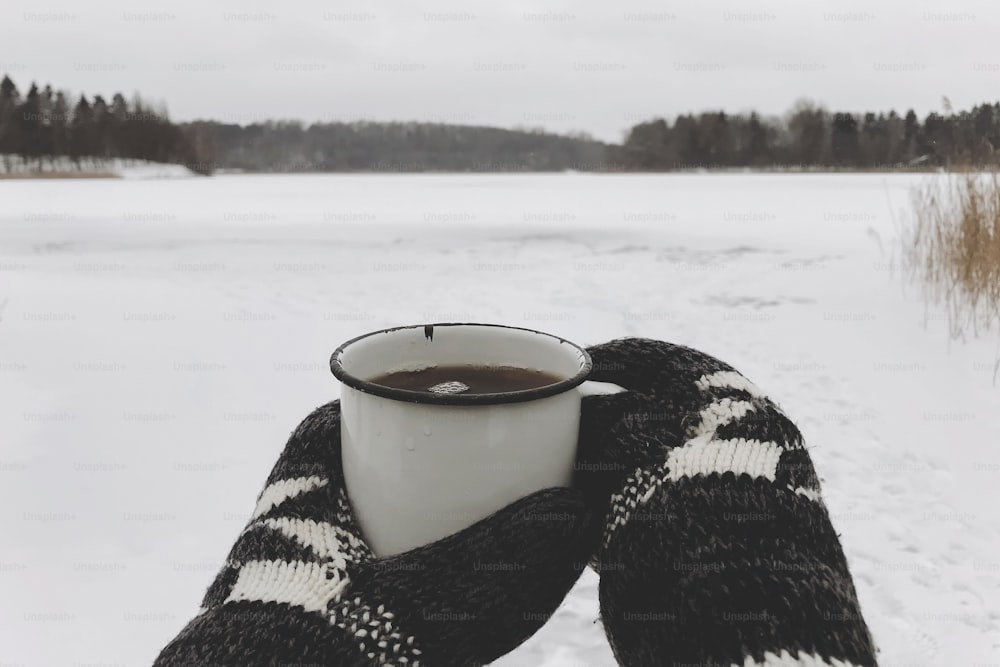 Hands in cozy gloves holding warm cup of tea on background of  snow lake in winter. Hiking and traveling in cold winter season. Warm drink. Space for text. Wanderlust