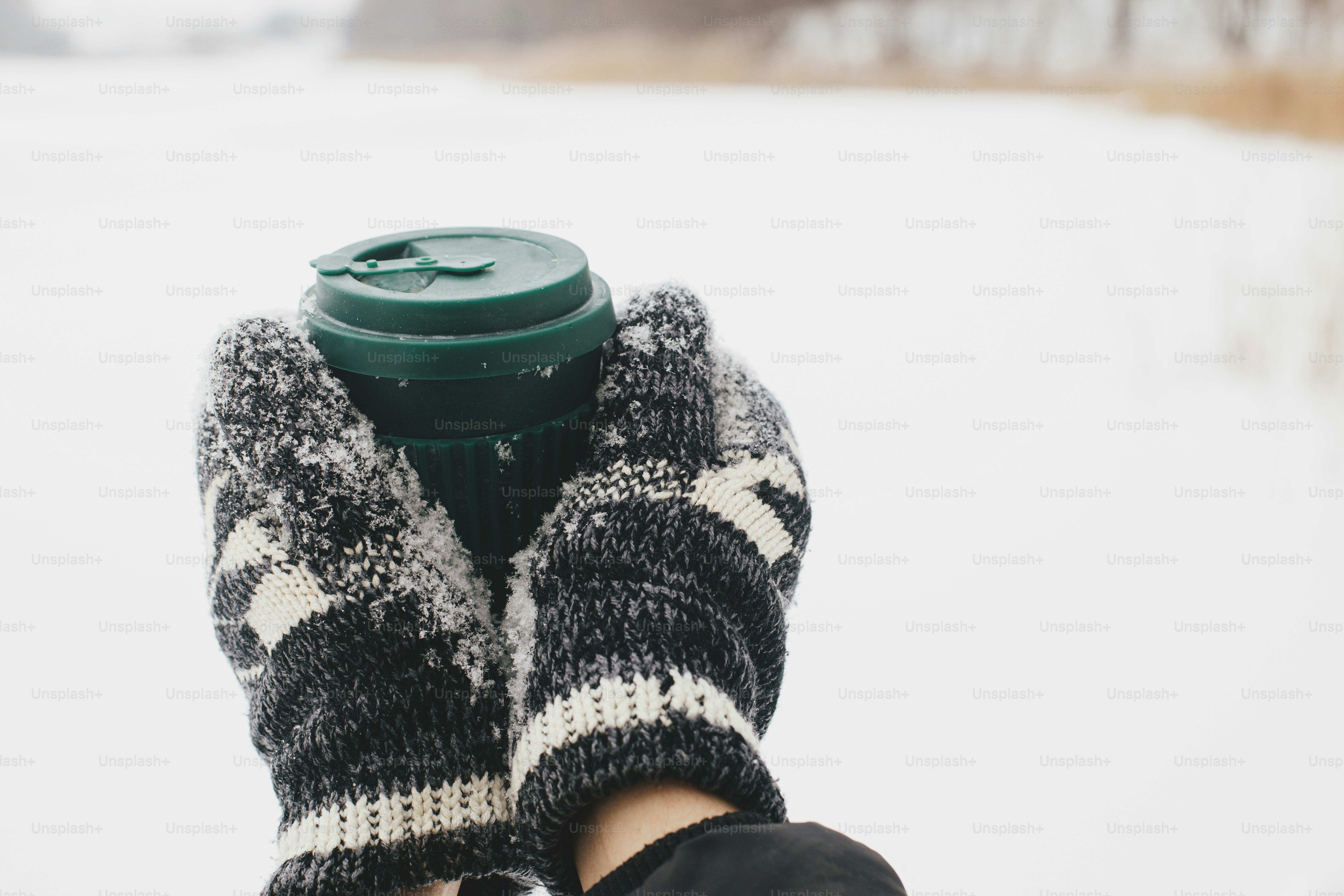 Hands in cozy gloves holding reusable warm cup of tea on background of snow lake in winter. Plastic free cup with warm drink. Hiking and traveling in cold winter season. Sustainable lifestyle
