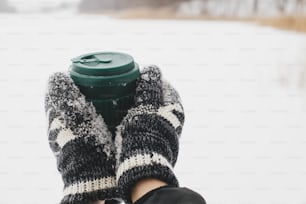 Hands in cozy gloves holding reusable warm cup of tea on background of snow lake in winter. Plastic free cup with warm drink. Hiking and traveling in cold winter season. Sustainable lifestyle