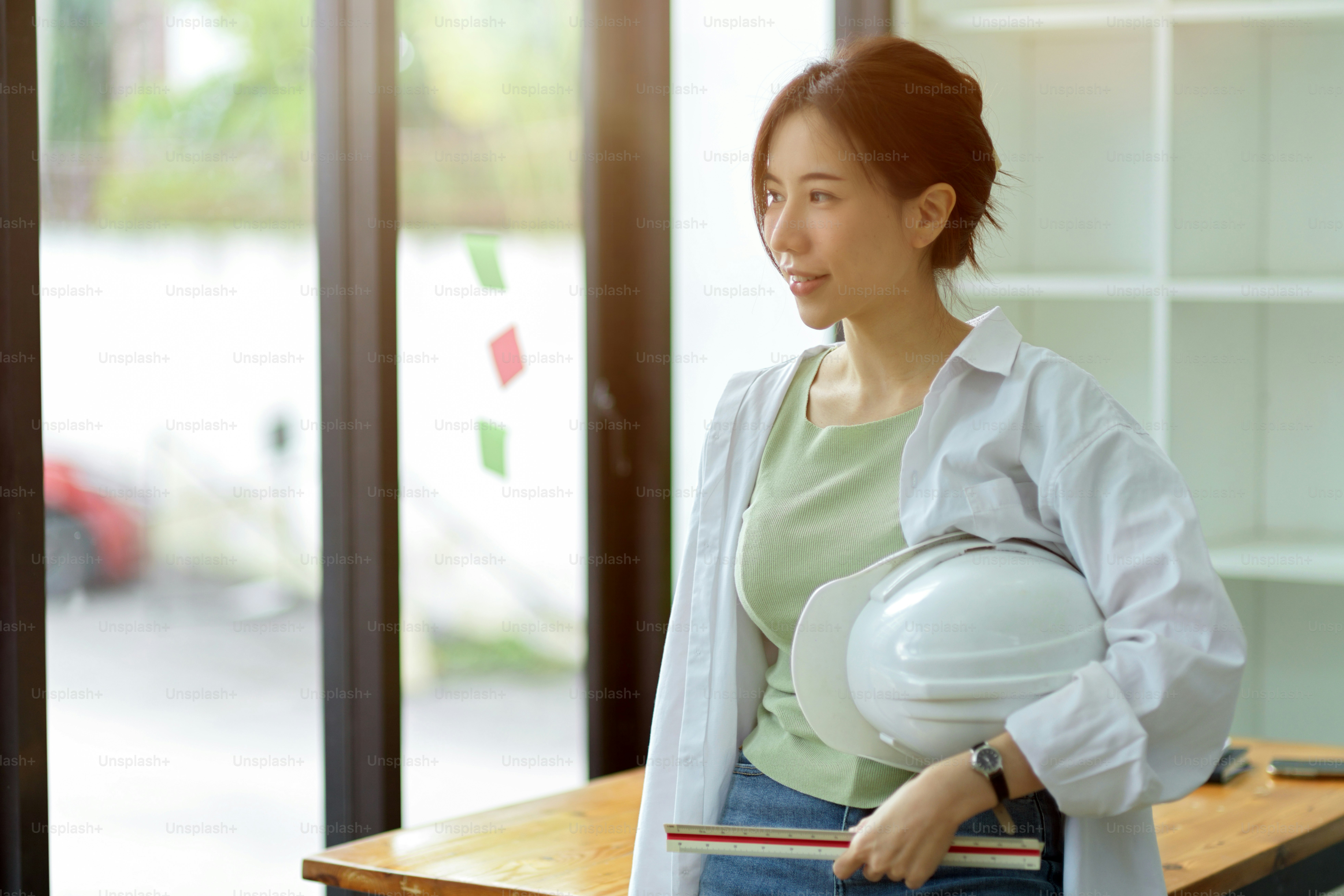Insinyur wanita muda melihat ke luar jendela, memegang penggaris dan topi keras di kantor insinyur