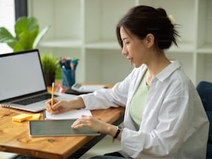 Hübsche Architektinnen, die einen digitalen Entwurf erstellen, einen Plan auf Notizblöcken und einem digitalen Tablet skizzieren, ein Laptop-Mockup in einem modernen Büroarbeitsplatz erstellen