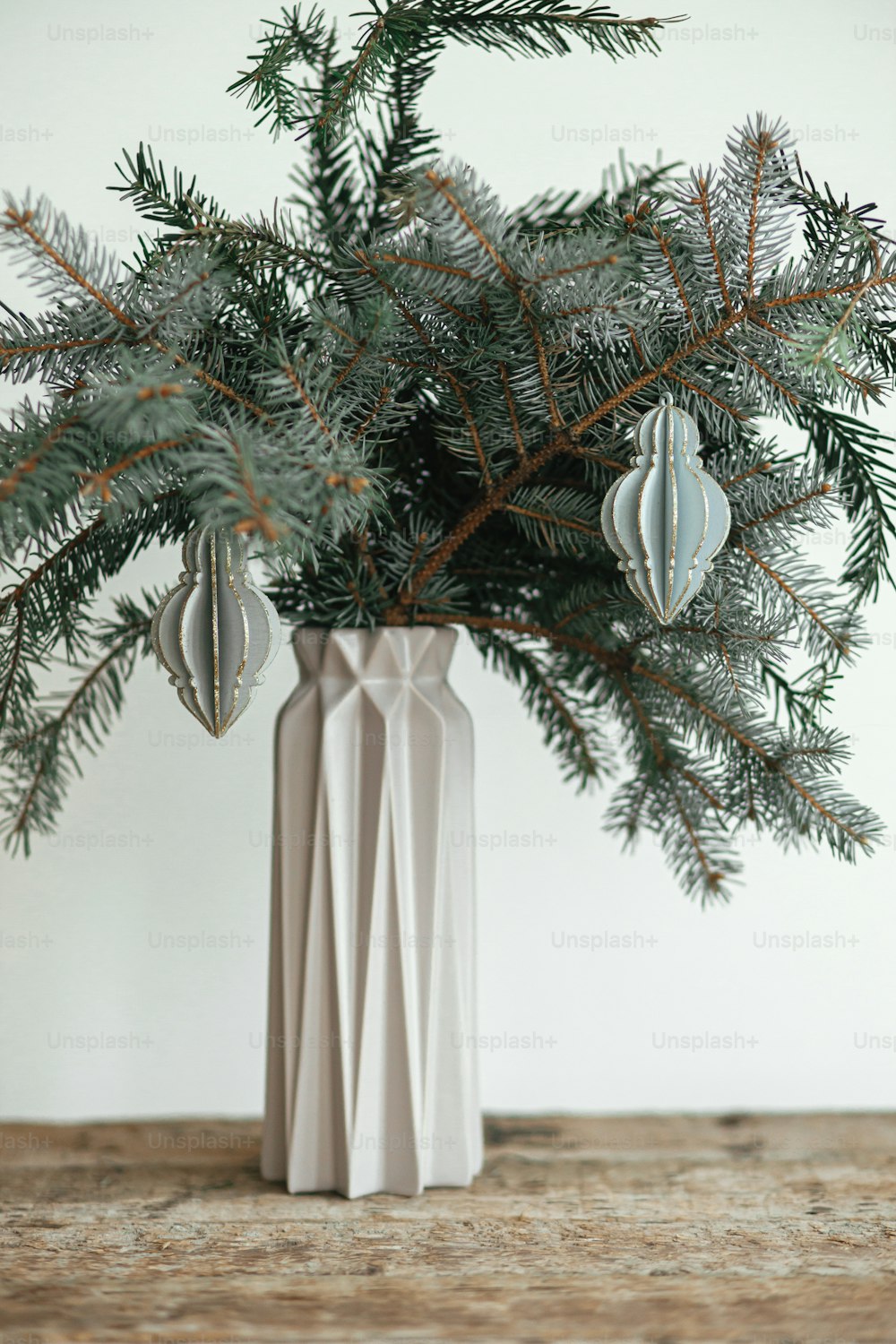 Stylish christmas ornament on fir branch in modern vase close up. Modern wooden bauble on branch on white background. Festive simple and creative decor in scandinavian room. Merry Christmas!