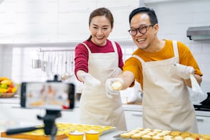 Asian couple bakery shop owner using smartphone with internet vlogging sweet dessert baking on social media together in the kitchen. Small business entrepreneur and online cooking class concept