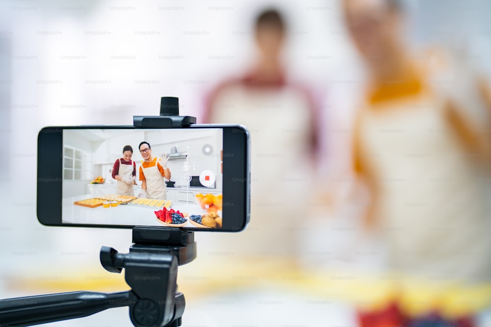 Asian couple bakery shop owner using smartphone with internet vlogging sweet dessert baking on social media together in the kitchen. Small business entrepreneur and online cooking class concept