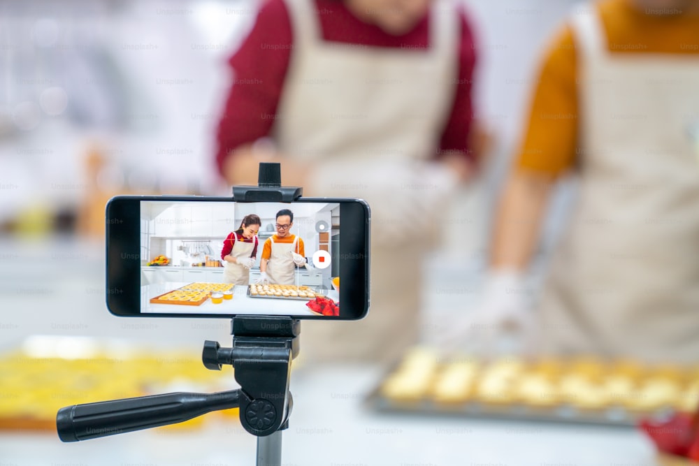 아시아 커플 베이커리 가게 주인은 인터넷 브이로깅으로 스마트폰을 사용하여 부엌에서 함께 소셜 미디어에서 달콤한 디저트를 굽고 있습니다. 중소 기업 기업가 및 온라인 요리 수업 개념