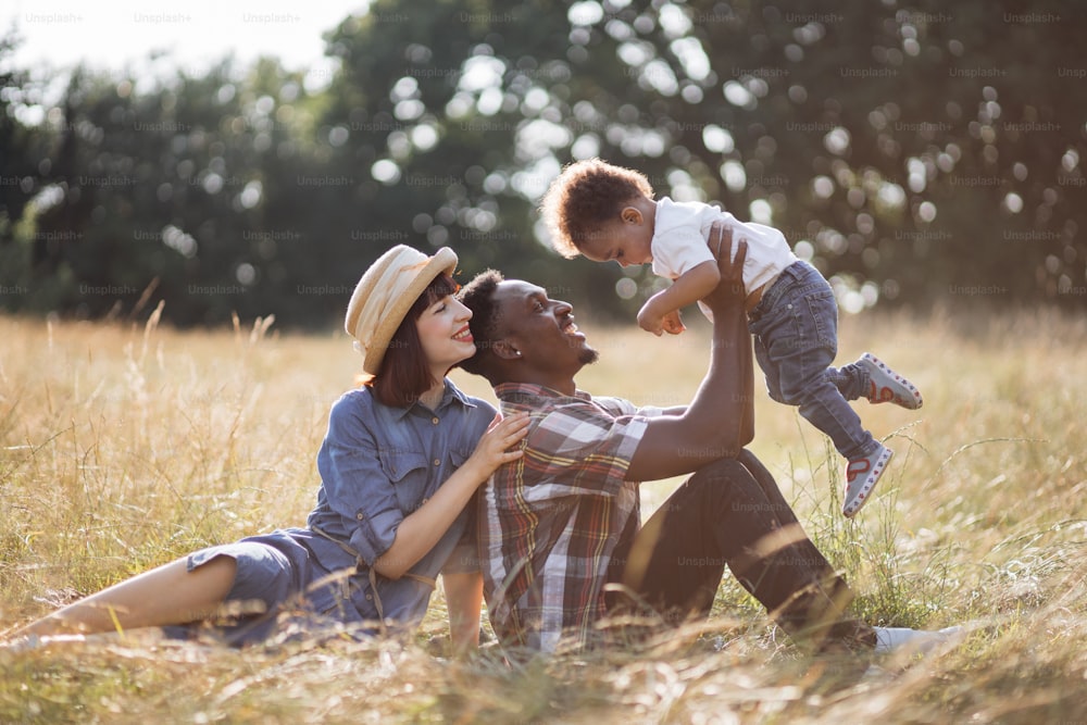 Positive multi ethnic family of three in casual wear playing together during sunny day outdoors. Caucasian woman hugging her african husband that lifting little son on hands.