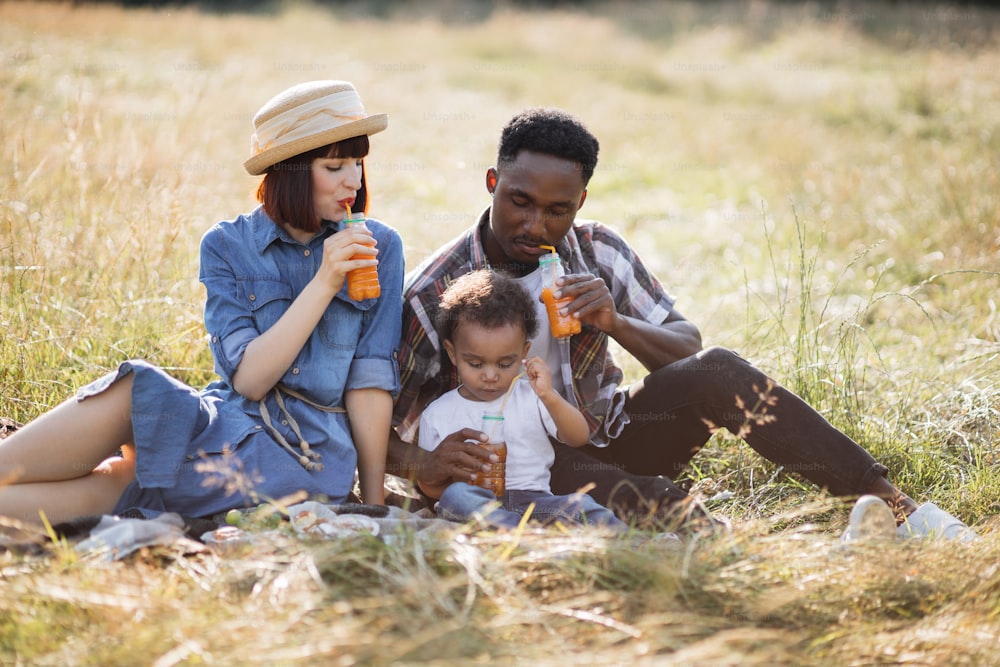 Positive young parents and their cute son sitting together on grass and drinking fresh juice from straw. Adorable multi ethnic family enjoying picnic time during summer days.