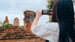 タイのアユタヤで休暇旅行を過ごしながら写真を撮るためにスマートフォンを使用している旅行者のアジア人女性、伝統的な都市の素晴らしいランドマークで彼女の旅を楽しむ日本人女性観光客。