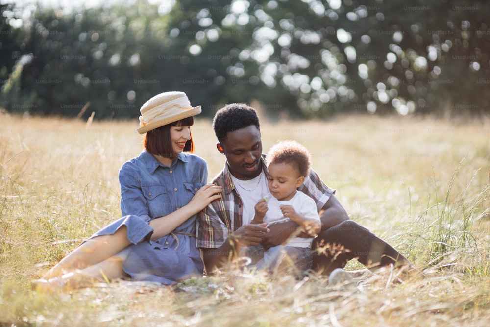 Multiracial parents and their cute little son having summer picnic on fresh air. Happy family of three, chatting, smiling and eating fruits outdoors. Relaxation concept.