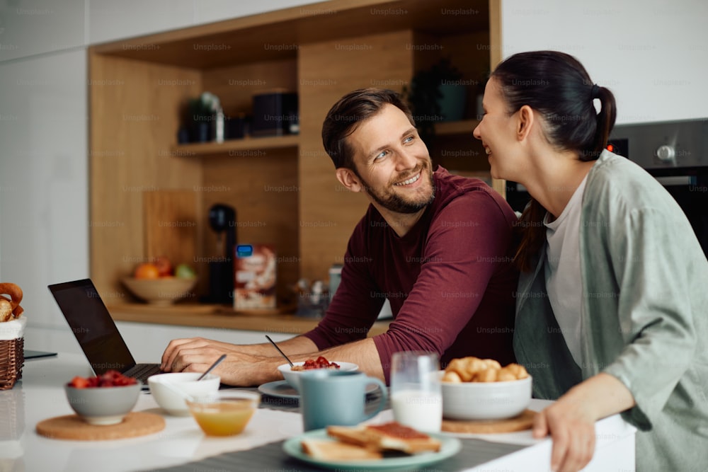 Young happy man communicating with his wife while using laptop during their breakfast at home.
