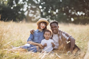Pai afro-americano e mãe caucasiana desfrutando de piquenique de verão com seu filho pequeno bonito. Linda família sentada na grama e sorrindo sinceramente. Conceito de relaxamento e amor.