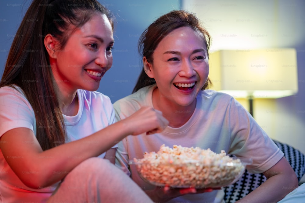 Millennial Asian woman friends sitting on sofa watching movie on tv with eating popcorn snack together in living room. Female friendship enjoy and having fun weekend activity lifestyle with home entertainment.