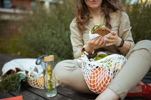 街の屋外に座っている間、ストリングバッグ、水筒、サンドイッチを食べている全体的な女性のクローズアップ。