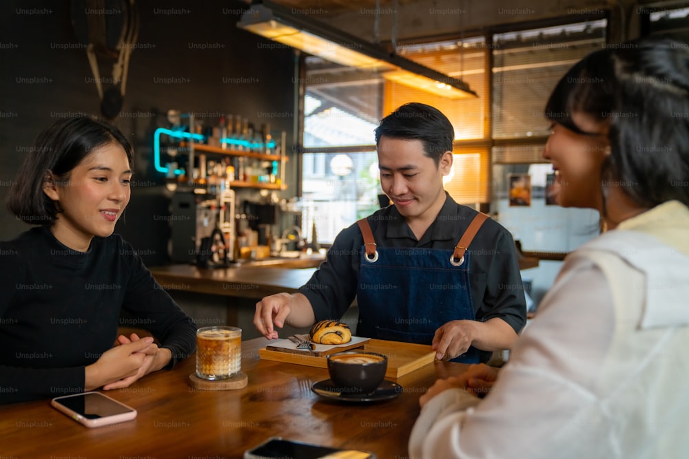 Asian man coffee shop waiter barista serving coffee latte and bakery to woman customer on the table at cafe. Attractive female friend meeting and talking together at restaurant. Small business concept.