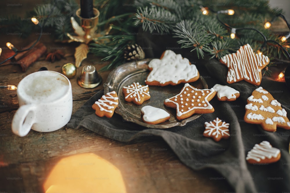 Christmas gingerbread cookies, coffee in stylish white cup, fir branches, warm lights on napkin and rustic wooden table. Cozy atmospheric image. Winter hygge. Happy Holidays!